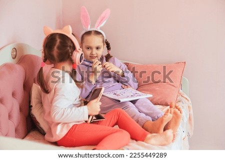 two sister girls have fun at home, draw a bunny and listen to music in headphones, the older girl has a hairband with rabbit ears on her head, waiting for the Easter holiday