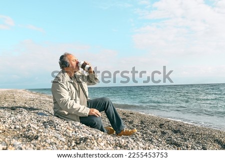 Happy middle-aged bearded man in listening music or podcast from smartphone application in headphones and drinking coffee while sitting on winter on spring beach. Relaxing outdoors.