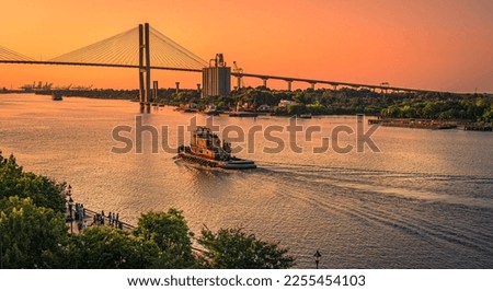 Artistic view of evening in Savannah, Georgia, as tourists gather on the river bank and a tug boat makes a wake heading into the golden sky under the Talmadge Memorial Bridge. Royalty-Free Stock Photo #2255454103