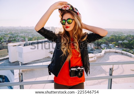 Outdoor fashion portrait of stylish photographer girl holding vintage retro camera, wearing bright swag hat, trendy sunglasses and leather jacket, amazing view of city from the roof.