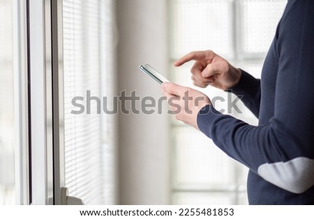 Man standing next to windows at home and using smartphone. Close up of a man in sweater standing in front of windows at home and holding smartphone pointing on the screen with the finger