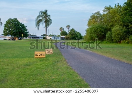 Two yellow wooden signs, no carts and cart path only this hole, on green grass next to a paved cart path on a golf course. It's a sunny summer's day with palm trees, woods and trailers along the edge.