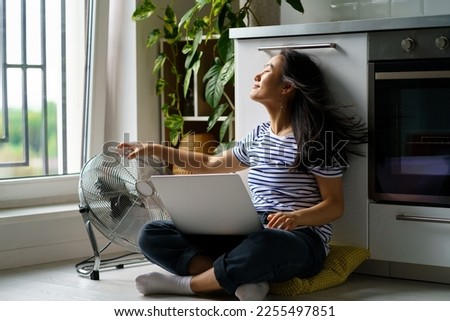 Satisfied Asian woman sits on floor with laptop on crossed knees, enjoying wind from electric fan. Relaxed Korean female freelancer takes break from work, enjoys the fresh breeze from fan. Summertime Royalty-Free Stock Photo #2255497851