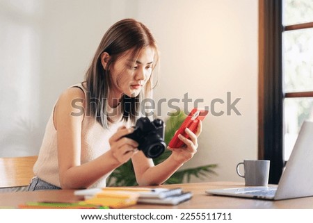 Female fashion artist holding camera and smartphone to checking photo of new fashion collection while working on laptop and designing new fashion clothes in workshop studio.