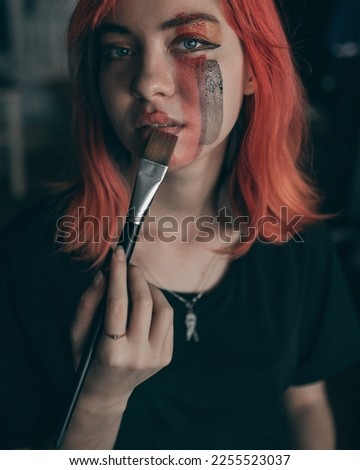 Young redhead woman artist applies red and black watercolor paint to her face with brush