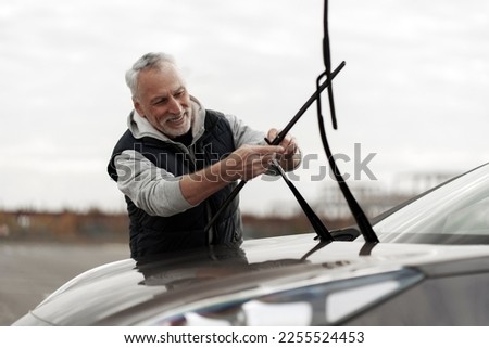 Handsome smiling senior Caucasian man, driver installing new windshield wipers by himself at outdoors parking lot. Automobile industry. Safety driving. Car maintenance. People. Auto service concept Royalty-Free Stock Photo #2255524453