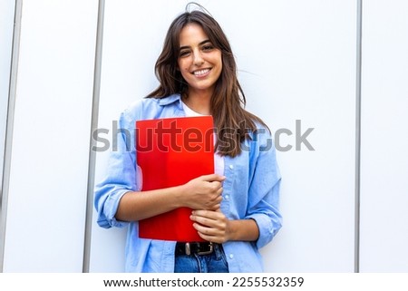Happy, smiling female college student looking at camera holding red folder.