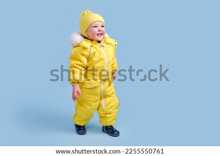 Happy toddler baby in winter clothes snowsuit on studio blue background. A child in a warm yellow jumpsuit with a hood. Kid aged one year five months Royalty-Free Stock Photo #2255550761