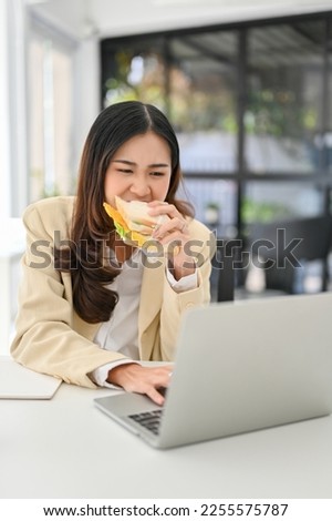 Busy and hardworking millennial Asian businesswoman eating a sandwich while working on her project on laptop in the office. deadline, multiple tasks Royalty-Free Stock Photo #2255575787