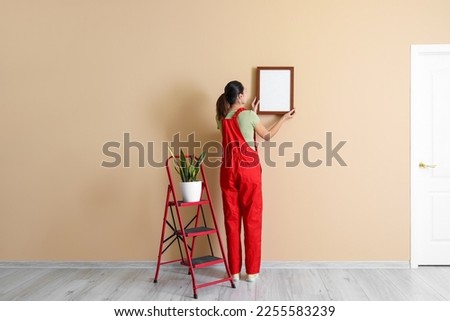 Young woman hanging frame on beige wall, back view