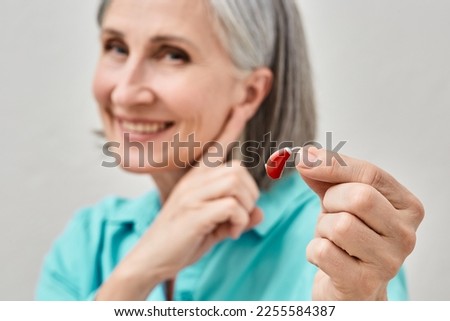 Senior woman holding hearing aid in hand on foreground and pointing finger at her ear, soft focus. Treatment of deafness in people with hearing aids Royalty-Free Stock Photo #2255584387