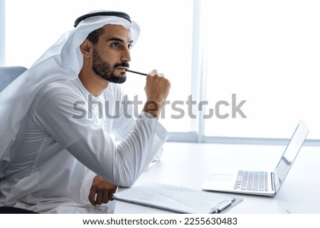 Handsome man with dish dasha working in his business office of Dubai. Portraits of a successful businessman in traditional emirates white dress.  Royalty-Free Stock Photo #2255630483