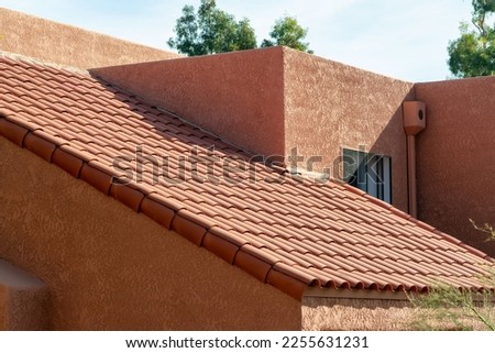 Long slanted adobe style roof with red tiles and orange stucco exterior with back and front yard trees in a neighborhood. In suburban area in late afternoon sun with desert design and facade. Royalty-Free Stock Photo #2255631231