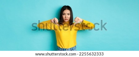 Disappointed asian woman frowning upset, showing thumbs down in dislike and disapproval, standing over blue background, staring at camera.