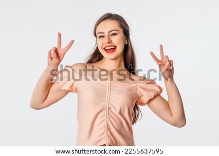 Positive charming brunette girl shows peace sign, makes victory gesture, having fun, posing against white studio background