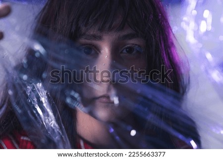 Portrait of white teenage girl in creative makeup looking at camera with serious expression through layer of plastic wrap. Artistic photography. Horizontal shot. High quality photo