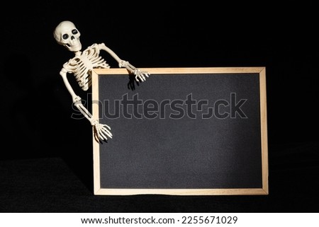 Funny human skeleton holding an empty black blank on a black background, copy space, mockup. Halloween Greeting Card