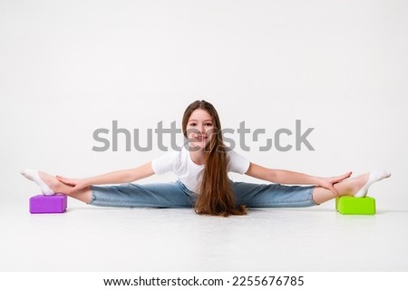 A teenage girl in jeans and a white t-shirt stretches her legs with green and purple styrofoam cubes. White background. The concept of sports, yoga.