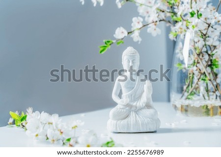 Decorative white Buddha statuette with blooming tree branches in the vase on the light background. Meditation and relaxation ritual. Buddha birthday. Selective focus