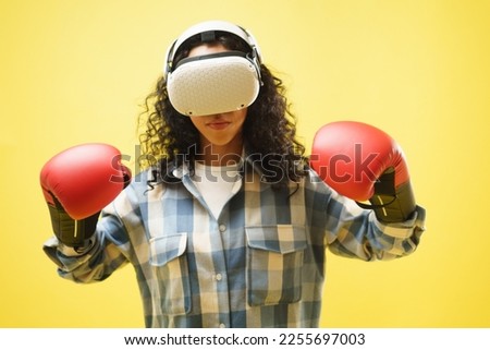 Portrait of young woman wearing VR goggle and boxing gloves holding hands up in a guard position, enjoying boxing game, gamer, hobby over yellow background. 