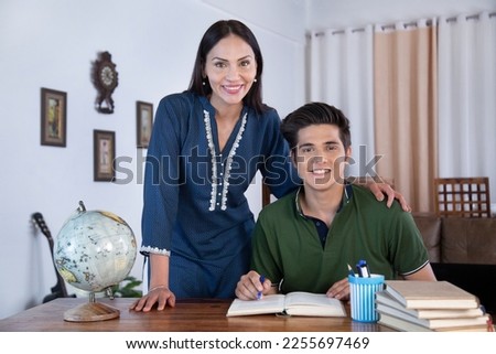 Photo shot of Indian teenager son and young mother both are smiling together. Indian teenager son and young mother both are smiling while doing homework at home, mummy standing near her seated son Royalty-Free Stock Photo #2255697469
