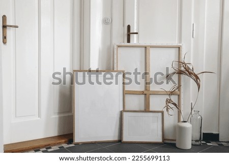 Boho wooden picture  frames set and blank canvas mockups on grey tiles floor. Artistic still life. Dry palm leaf in vase. Elegant minimal interior design, white wall and old doors background. Home.