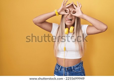 Young blonde woman standing over yellow background wearing headphones doing ok gesture like binoculars sticking tongue out, eyes looking through fingers. crazy expression. 