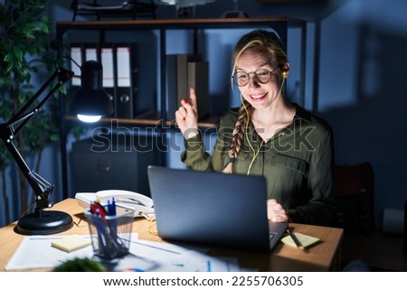 Young blonde woman working at the office at night with a big smile on face, pointing with hand and finger to the side looking at the camera. 
