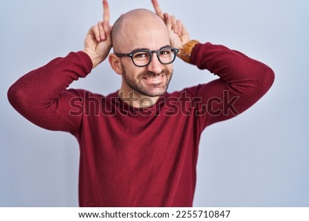 Young bald man with beard standing over white background wearing glasses posing funny and crazy with fingers on head as bunny ears, smiling cheerful 