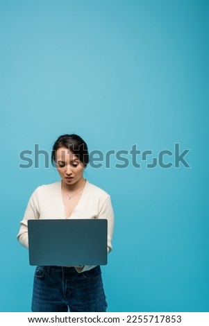 Focused asian freelancer using laptop isolated on blue