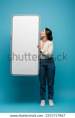 Side view of young asian woman pouting lips and holding big smartphone model on blue background Royalty-Free Stock Photo #2255717867
