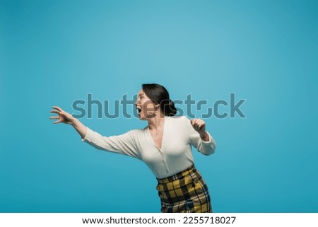 cheerful asian woman in plaid skirt smiling while gesturing isolated on blue Royalty-Free Stock Photo #2255718027