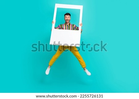 Full body size photo of funky jumping air trampoline man overjoyed wear striped shirt hold paper frame good mood isolated on cyan color background