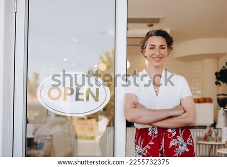 Successful cafe owner smiling at the camera while standing next to an open sign at the doorway of her restaurant. Female small business owner welcoming customers to her newly opened cafe