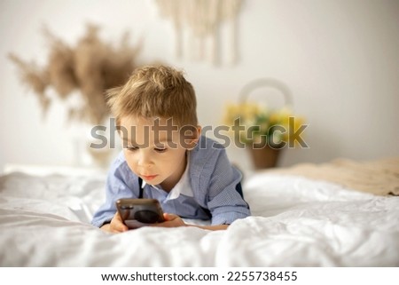 Blond preschool child, cute boy, playing on mobile phone in bed on a sunny day