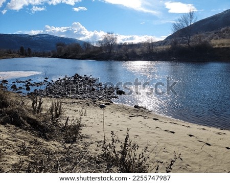 A beautiful photo of the beach. sun reflects off the water. sandy shores in the foreground and mountains in the distance with blue cloudy skies. 