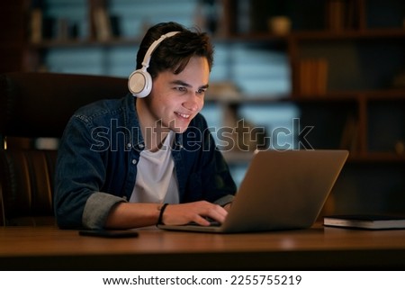 Deadline concept. Young smiling man networking and connecting to internet using a laptop late at night, he is sitting at desk and wearing wireless headphones at empty dark office Royalty-Free Stock Photo #2255755219