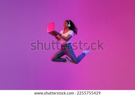Excited young black woman using laptop and jumping up in neon light, cheerful african american female wearing wireless headphones flying in air with modern portable computer over purple background