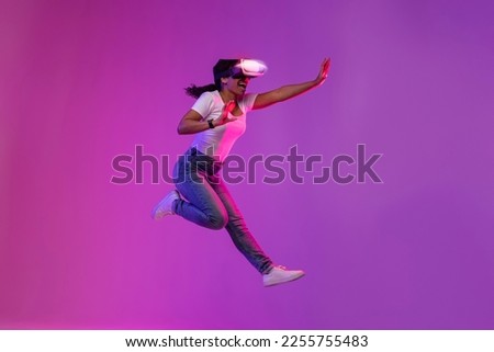 Excited Young Black Female Wearing Glowing VR Glasses Jumping In Neon Light, Amazed African American Woman Experiencing Virtual Reality And Having Fun Over Purple Studio Background, Copy Space