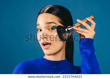 Woman with bold eyeshadow blends foundation on her face using a makeup brush. Flawless woman looking at the camera with an open mouth as she applies makeup on her face during her beauty routine. Royalty-Free Stock Photo #2255764621