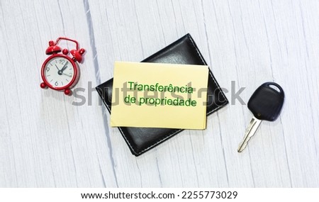 The phrase transfer of transfer of ownership in Brazilian Portuguese written on a piece of paper that is on top of a document holder. An alarm clock and a car key in the composition.
