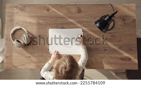 little blonde girl starts drawing with black marker, top down view of child at work desk at home or in kindergarten. Kid draws doodles on white blank sheet.