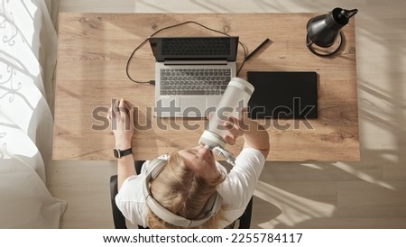 Top down view of girl at desktop with laptop and graphics tablet. Young blonde girl draws sketches or illustrations, in headphones and drink water from her bottle. Freelance work during day room.