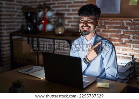 Young hispanic man with beard working at the office at night cheerful with a smile of face pointing with hand and finger up to the side with happy and natural expression on face 