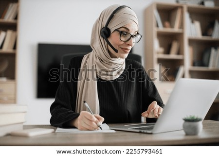 Attractive confident muslim business woman, office manager, wearing headset and hijab using laptop while making, writing financial report, using pen, on paper working indoors.