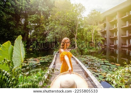 Happy woman in yellow dress dancing with straw hat in her hand, looking at camera. Summer vacation in resort. Female traveler on wooden bridge among tropical greenery and green pond with water lilies Royalty-Free Stock Photo #2255805533
