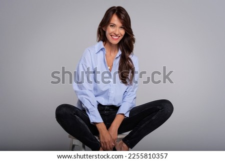Portrait of an attractive brunette woman wearing shirt and black trousers while sitting on stool at isolated grey background. Copy space.