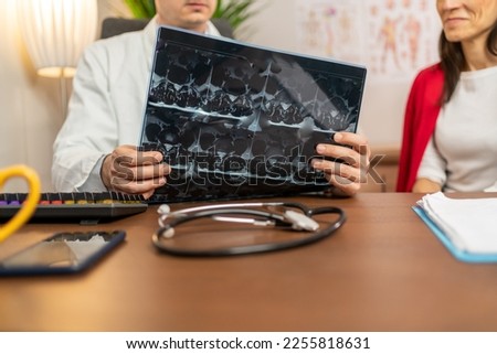 Serious man doctor and patient looking at the x-ray or MRI picture in hospital. Doctor attentively examines the MRI scan of the patient. Doctor examining MRI images of patient with multiple sclerosis  Royalty-Free Stock Photo #2255818631