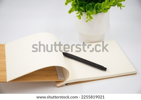 An open notepad with a black pen next to a green plant on a white background.