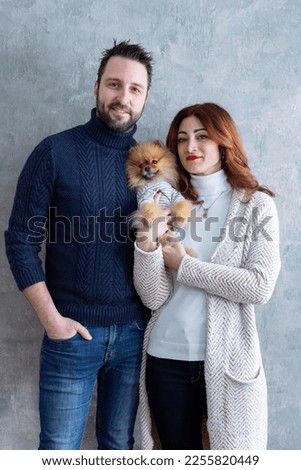 Selective focus vertical portrait of cute tall smiling guy in his thirties holding his red-haired girlfriend and Pomeranian dog close while standing against concrete background